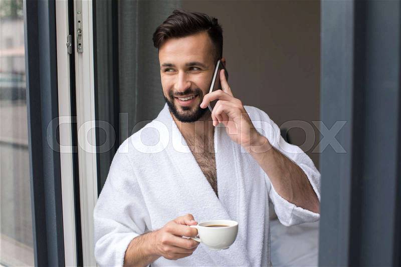 Handsome smiling bearded man in bathrobe talking on smartphone and drinking coffee at home , stock photo