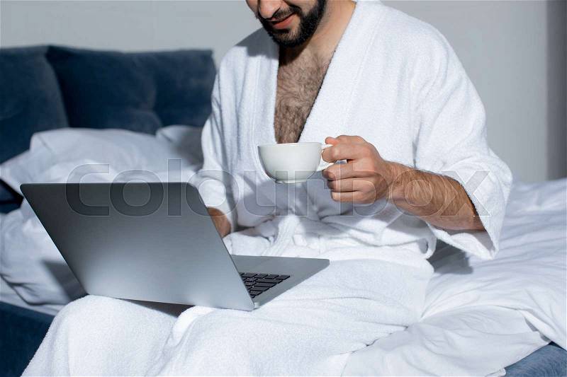 Cropped shot of bearded young man using laptop while drinking coffee in bed , stock photo