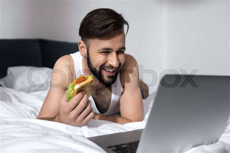 Bearded young man using laptop and eating sandwich while lying in bed at morning, stock photo