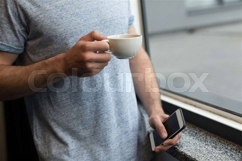 Cropped shot of young man using smartphone while drinking coffee in morning at home, stock photo