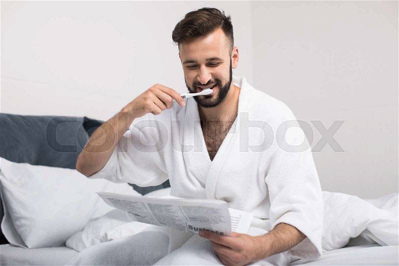 Handsome bearded man in bathrobe reading newspaper and brushing teeth while sitting on bed, stock photo