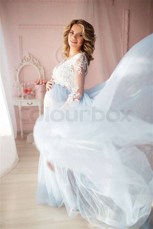 Pretty pregnant woman is wearing blue flying dress in studio with home interior, stock photo