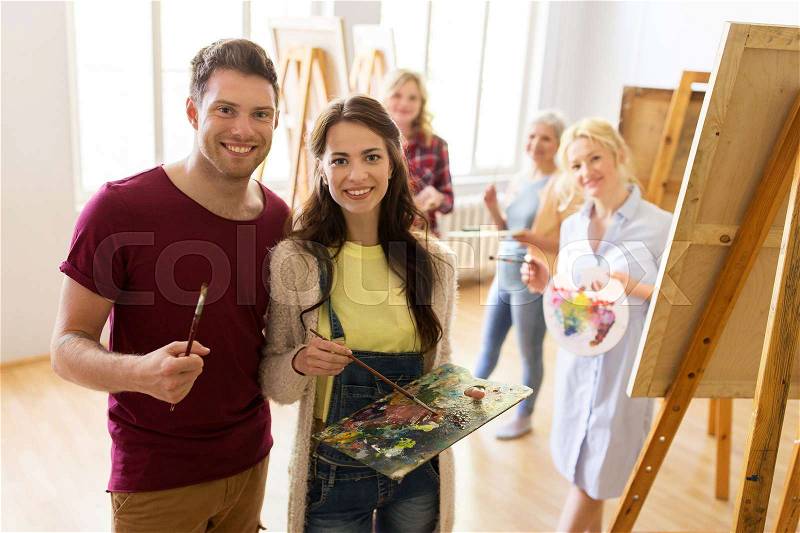 Creativity, education and people concept - group of artists or students with brushes and palettes painting on easels at art school studio, stock photo