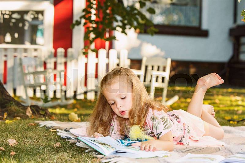 Cute little blond girl reading book outside on grass against green lawn, stock photo