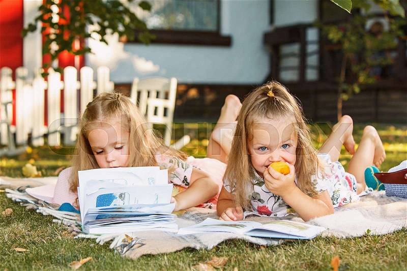 Cute little blond girls reading book outside on grass against green lawn, stock photo