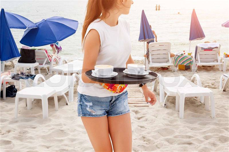 Girl waiter carrying coffee on the beach. Woman serving drinks. Sea and beach sunbeds on the background. Summer vacation at the sea, stock photo