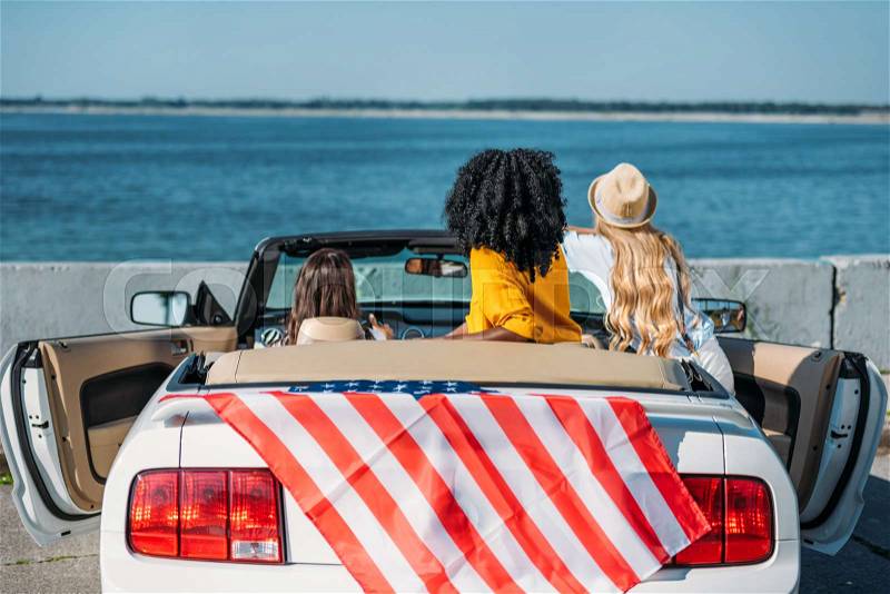 Back view of friends sitting in car with american flag at seaside, stock photo