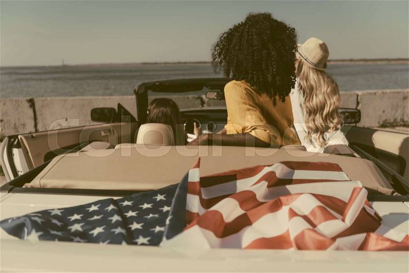 Back view of friends sitting in car with american flag at seaside, stock photo