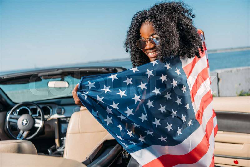 Back view of african american woman with american flag sitting in car at seaside, stock photo