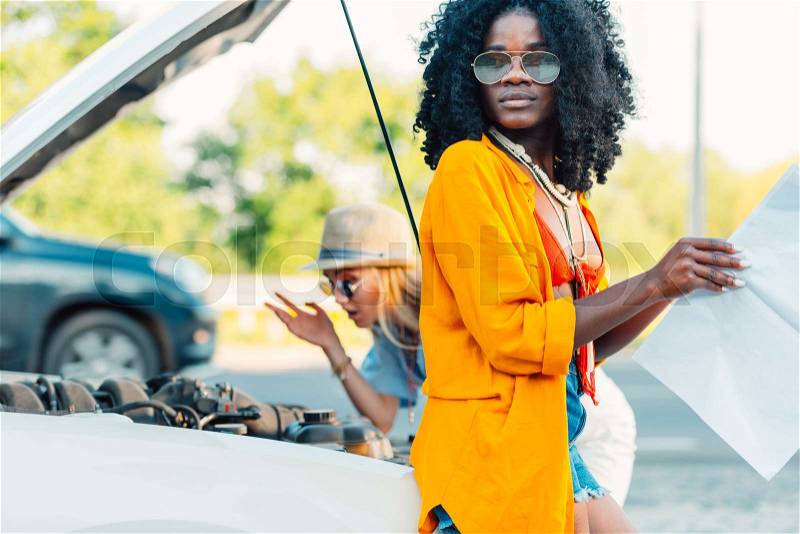 African american woman looking for destination on map while friend checking broken car, stock photo