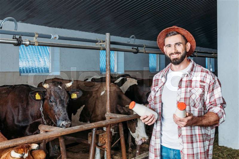 Handsome bearded farmer holding bottles with milk and smiling at camera in stall , stock photo
