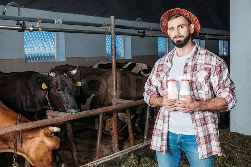 Handsome bearded farmer holding bottles with milk and smiling at camera in stall , stock photo