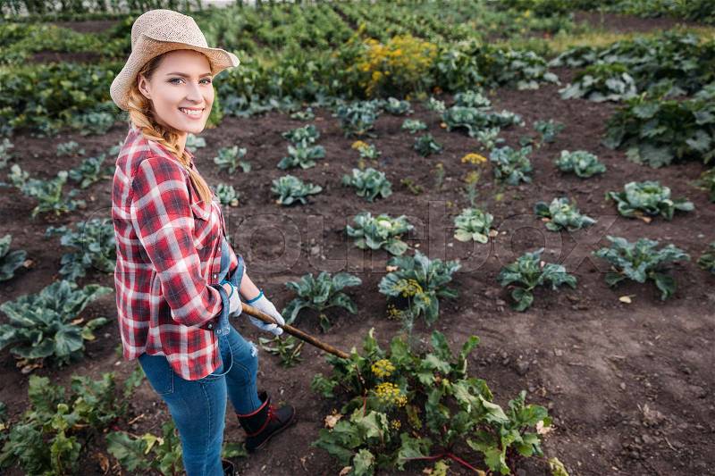 Beautiful young farmer in hat hoeing beets and smiling at camera on field, stock photo