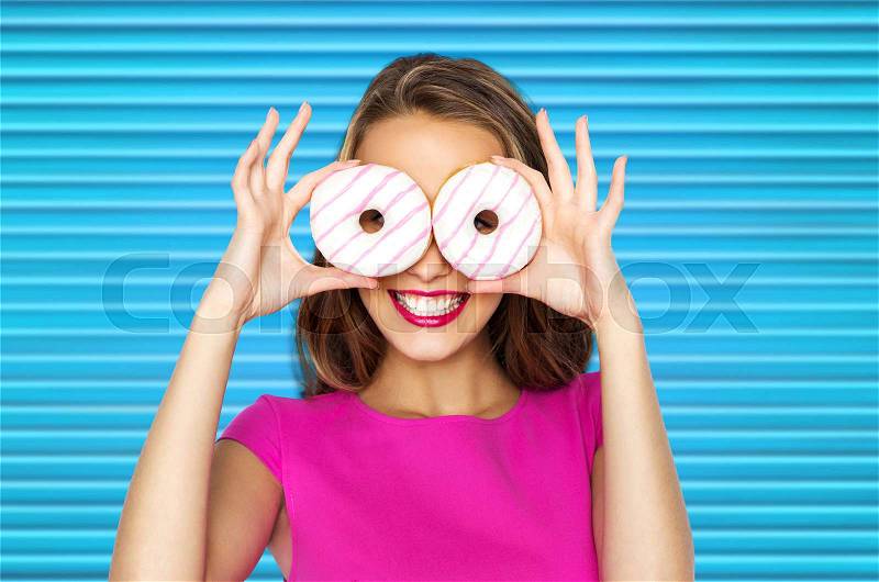 People, sweets and fast food concept - happy young woman or teen girl in pink dress having fun and looking through donuts over blue ribbed background, stock photo