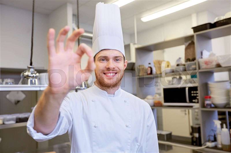 Cooking, profession and people concept - happy male chef cook in toque at restaurant kitchen showing ok hand sign, stock photo
