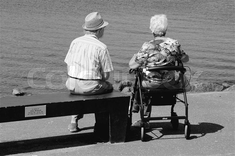 Retired couple, man is sitting on the bench and the woman on her rollator and have a beautiful view over Loch Lomond in Scotland in the summer in black and white, stock photo