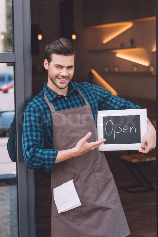 Portrait of smiling barista pointing at chalkboard with open word in hand, stock photo