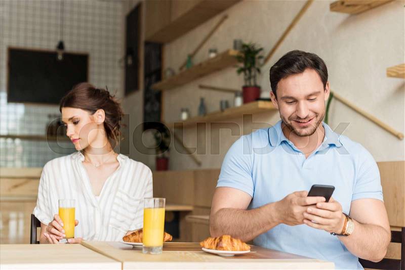 Addicted smiling man using his smartphone and unhappy woman sitting on background, stock photo