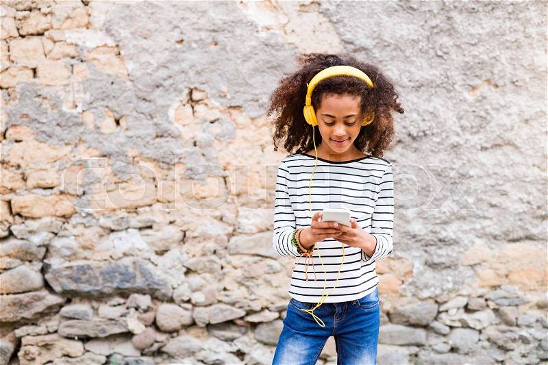 Beautiful african american girl with curly hair outdoors against stone wall, holding smart phone, wearing yellow headphones, listening music, stock photo
