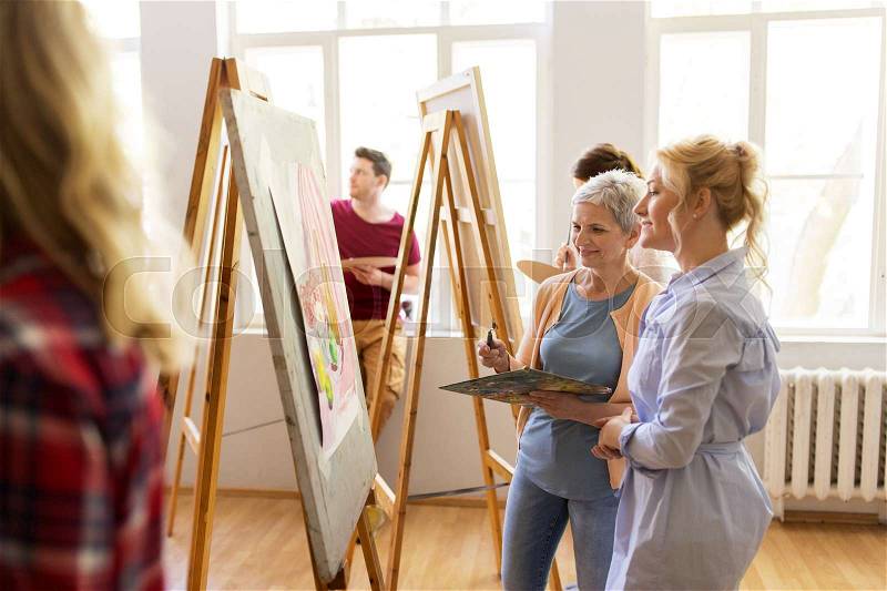 Creativity, education and people concept - women artists discussing painting on easel at art school studio, stock photo