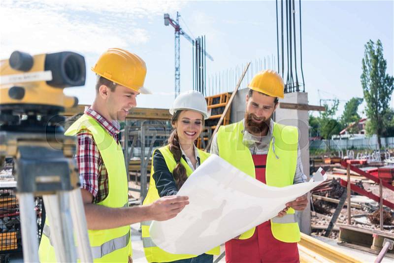 Young and confident female architect smiling while sharing ideas about the plan of a building under construction outdoors with two blue-collar workers, stock photo