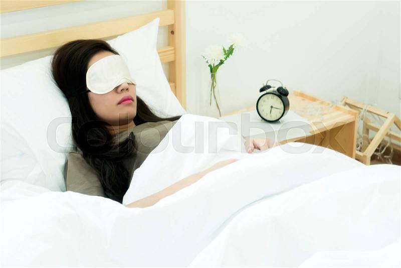 The young beautiful woman wearing eyes mask lying on white bed with comfortable sleeping, A clock show the morning time to wake up, stock photo