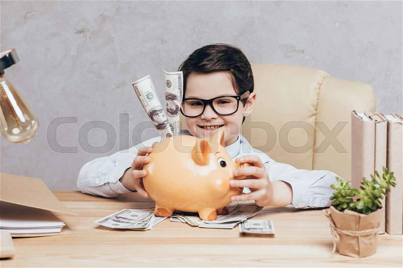 Smiling little boy sitting at workplace with piggy bank and cash isolated on grey, stock photo