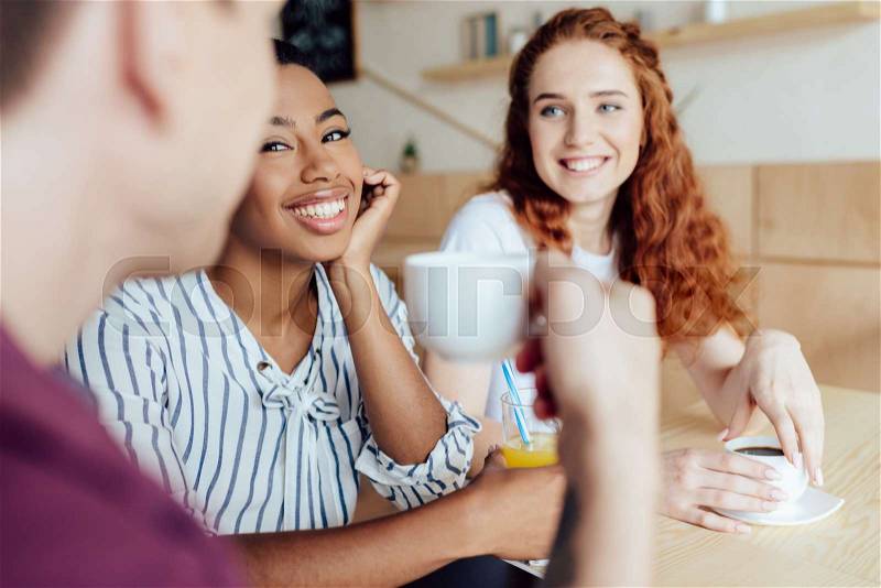 Smiling young multiethnic friends drinking coffee and juice while sitting together in cafe, stock photo