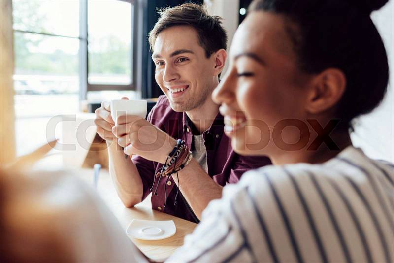 Smiling young multiethnic friends drinking coffee and looking away in cafe, stock photo