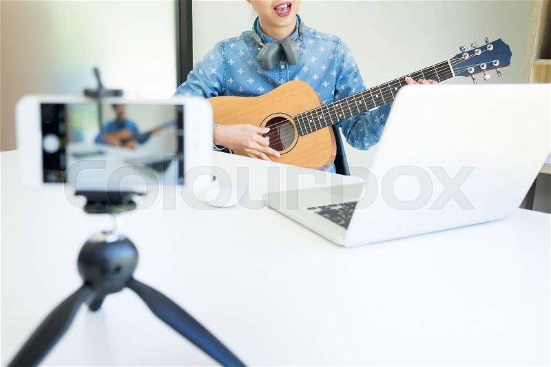 Women sing a song with guitar in hands use camera to broadcast live video to social network by internet at home, stock photo