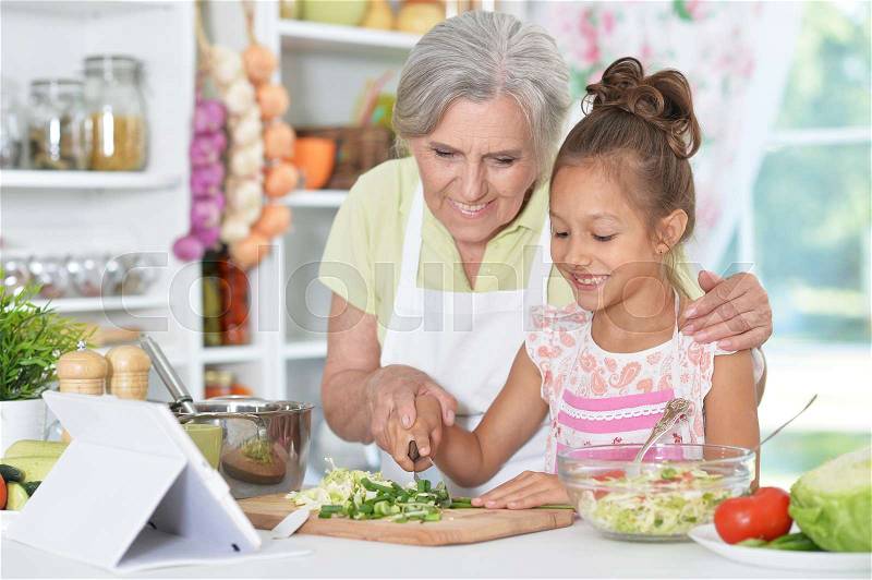 Grandmother and granddaughter preparing dinner on table with tablet, stock photo