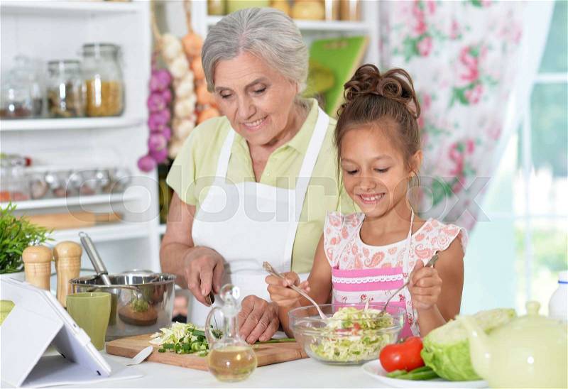 Grandmother and granddaughter preparing dinner on table with tablet, stock photo