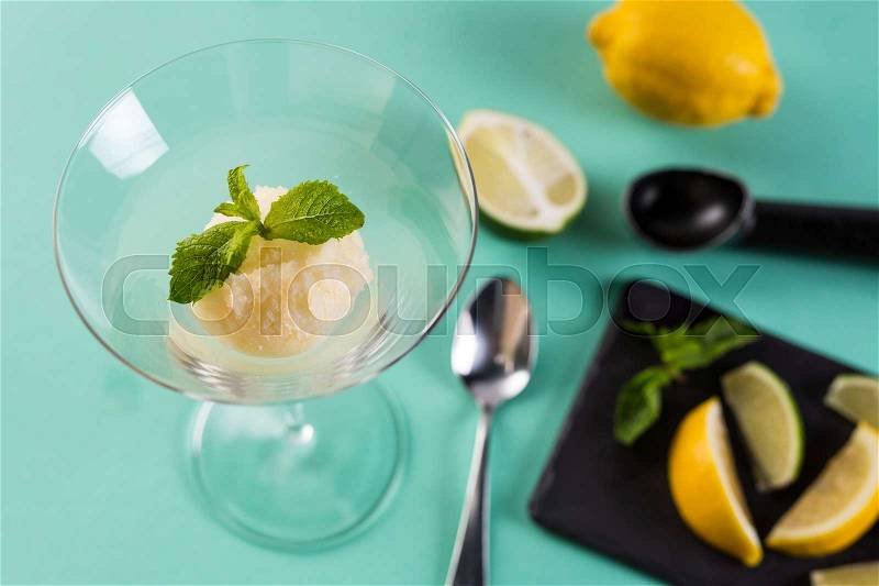 Lemon sorbet garnished with mint leaf. Tasty sorbet in high glass with spoon. Citrus ice-cream with lemon and lime on Turquoise background, stock photo