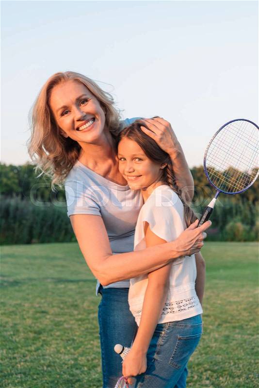 Portrait of smiling grandmother with badminton racket hugging granddaughter on meadow, stock photo