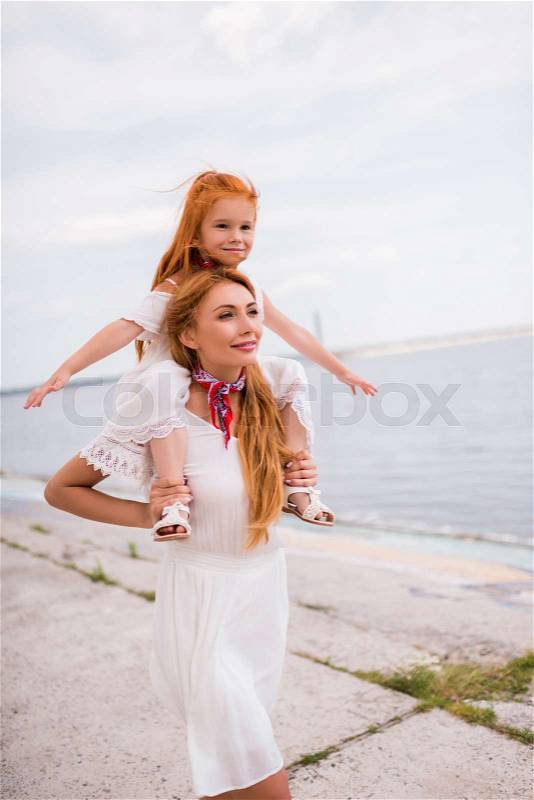 Happy young mother carrying adorable little daughter on neck while walking at seaside, stock photo