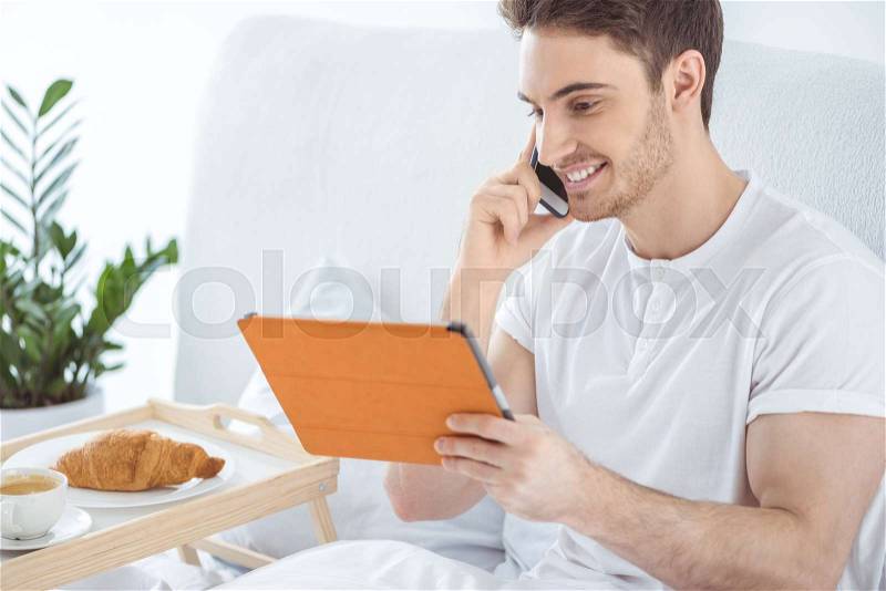 Man using digital tablet and smartphone while sitting in bed, stock photo