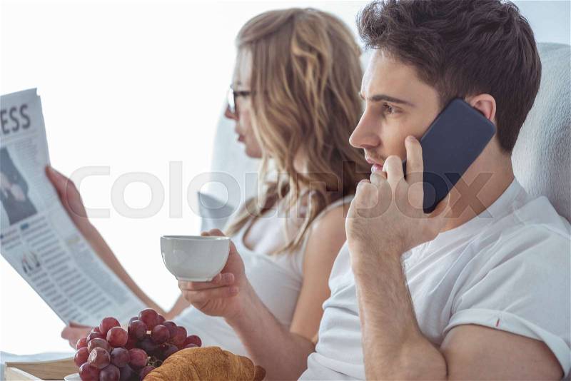 Young couple using smartphone and reading newspaper during breakfast in bed , stock photo