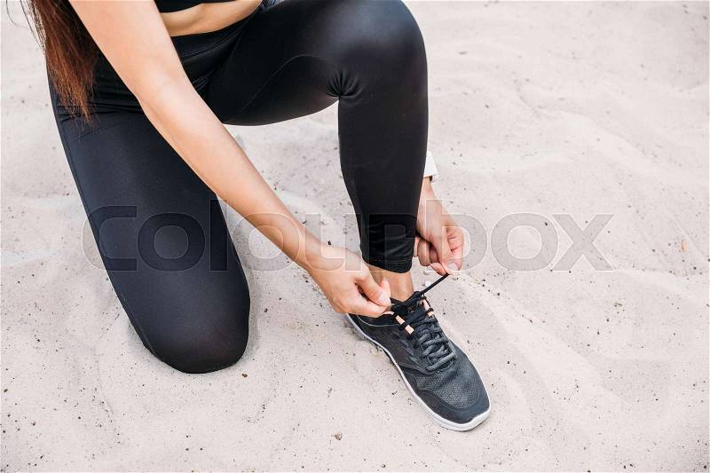 Cropped view of sportswoman in sportswear tying shoelaces on sand, stock photo