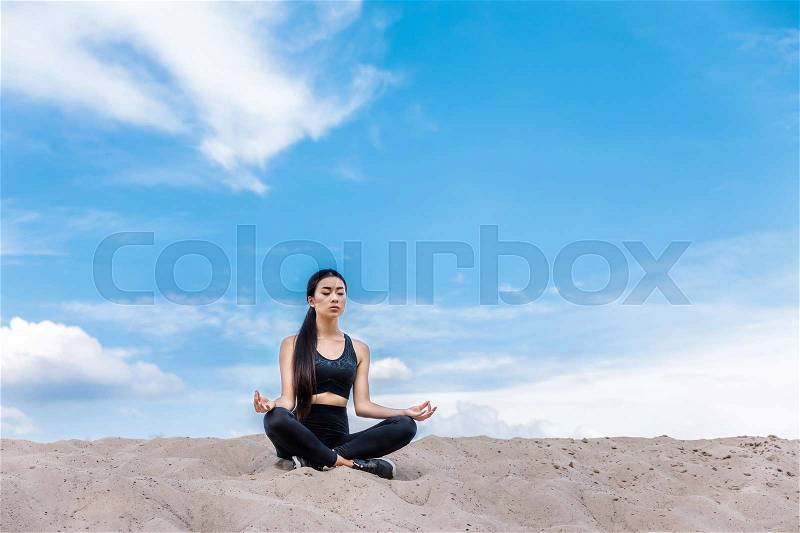 Asian woman meditating in lotus yoga pose on sand against cloudy sky , stock photo