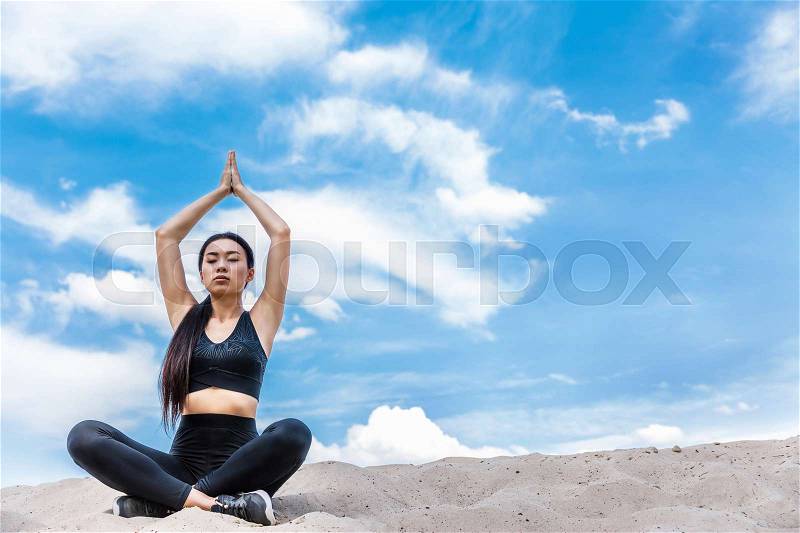 Asian woman meditating in lotus yoga pose with namaste mudra on sand against cloudy sky , stock photo