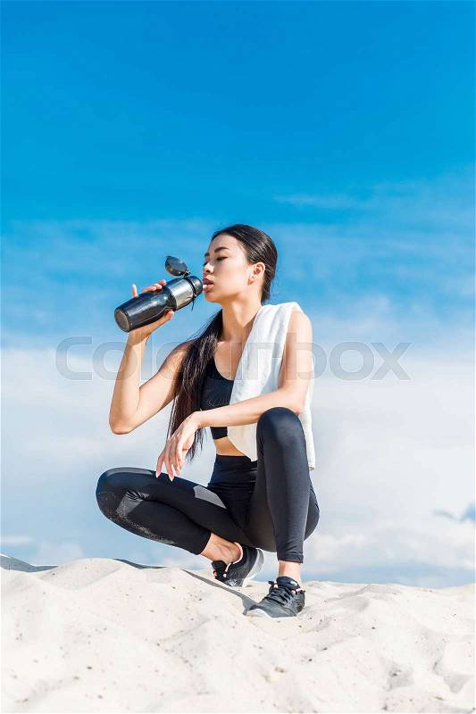 Tired asian sportswoman drinking water with sport bottle resting on sand against cloudy sky , stock photo