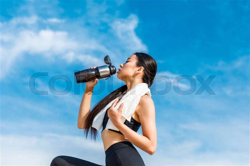 Tired asian sportswoman drinking water from sport bottle against cloudy sky , stock photo