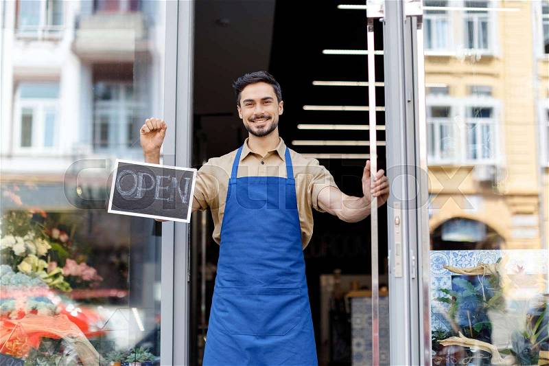 Happy young florist in apron holding open sign and smiling at camera, stock photo