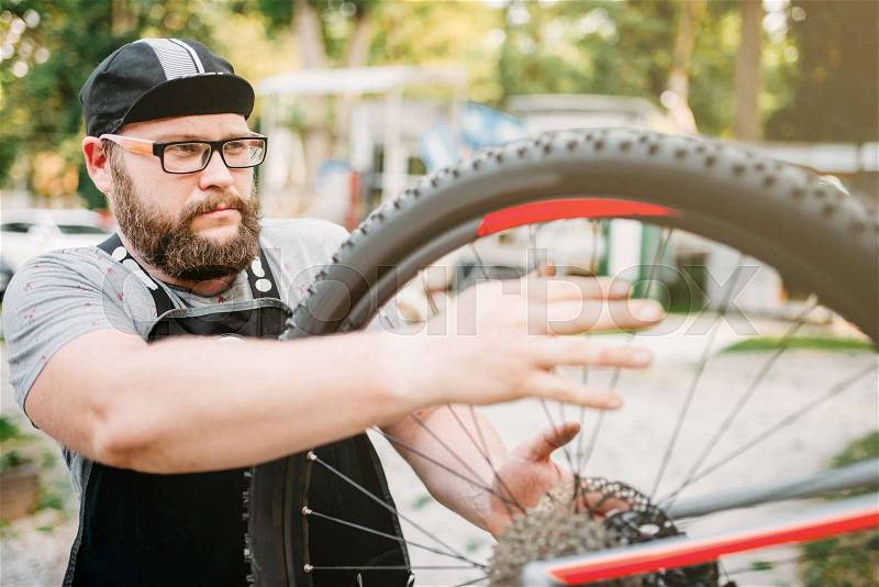 Bicycle repairman works with bike wheel, cycle workshop outdoor. Bearded mechanic in apron, stock photo