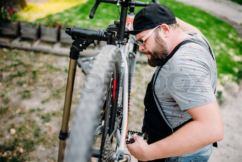 Repairman works with bike wheel, cycle workshop outdoor. Bearded bicycle mechanic in apron, stock photo