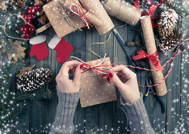 Christmas background. Top view of female hands wrap New Year present. Packed gifts and scrolls, spruce branches and tools on shabby wooden table. Workplace for preparing handmade decorations. Winter holidays concept, stock photo