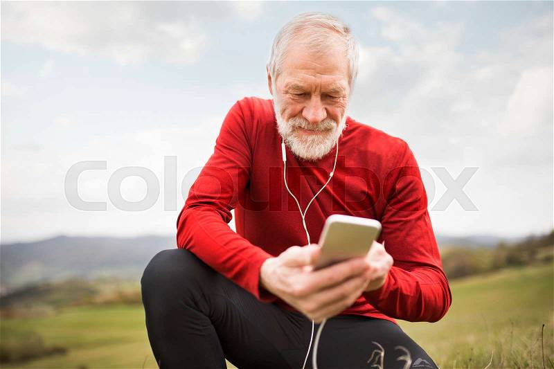 Active senior runner outside on green hills with smart phone and earphones, resting, listening music, stock photo