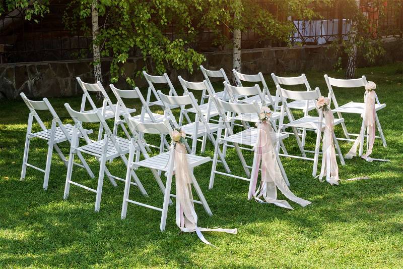 Beautiful wedding set up. Wedding ceremony in the garden. White wooden chairs decorated with flowers and pink ribbons standing in rows, stock photo