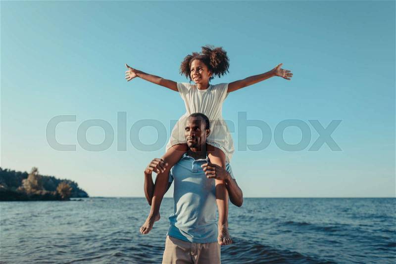 Happy african american father carrying adorable little daughter on neck at seaside, stock photo