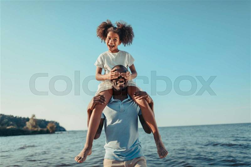 Smiling african american father carrying adorable little daughter on beach, stock photo